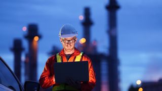 An industrial engineer standing outdoors, his face is lit by the light from a laptop and a power station blurred in the background. He is wearing a hard hat and high-vis jacket over an orange jumper.