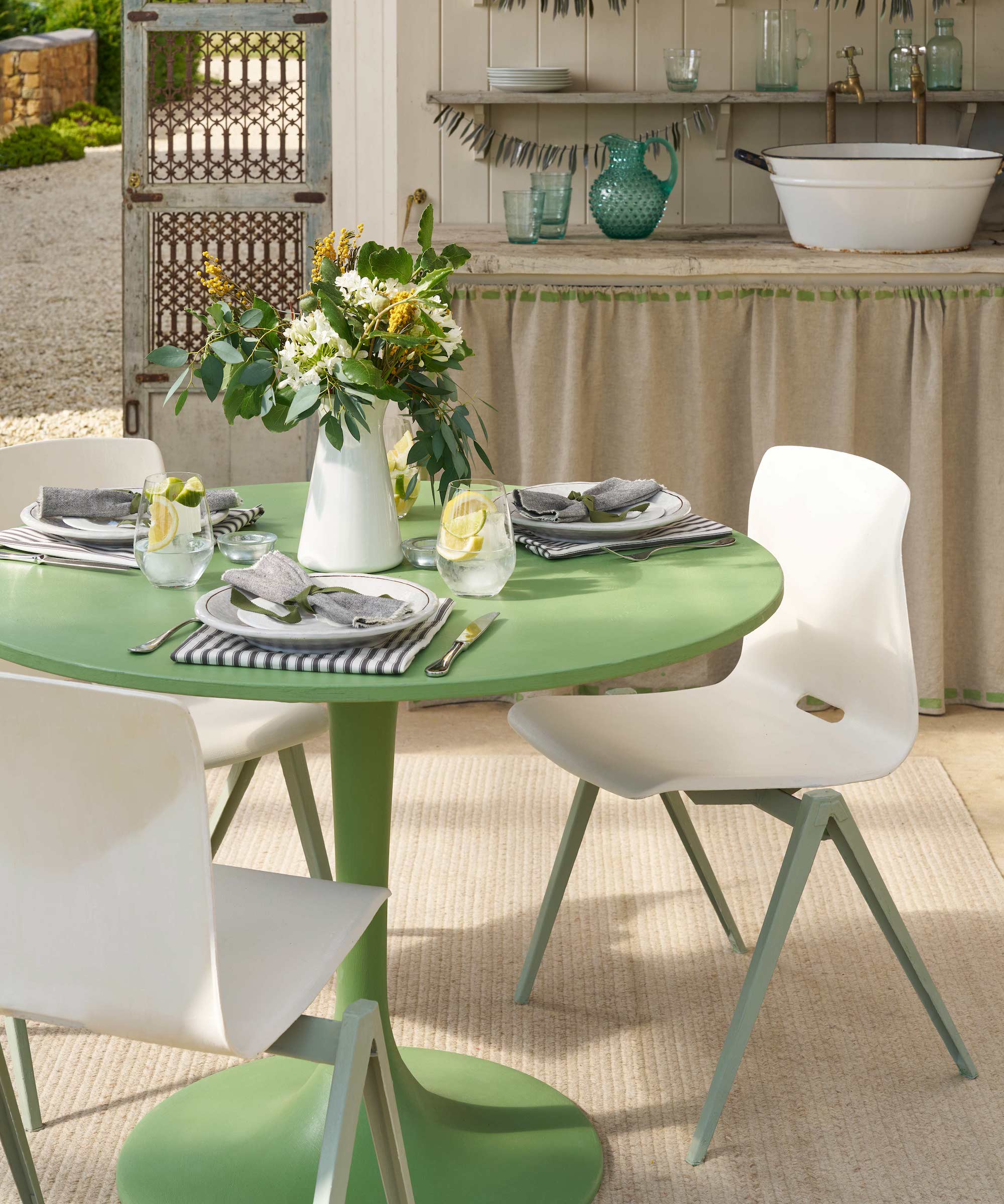 Round dining table painted with green Chalk Paint and white chairs with green legs with table setting shown in kitchen with skirted storage and wall shelves and door open to garden