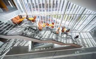 An interior view of the stairs and bottom floor captured from the floor above. Grey stairs with glass banisters and below that is a seating are with orange chairs and tables set against the glass panel windows