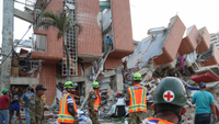 Buildings damaged by an earthquake, with people in helmets and high-vis jackets outside