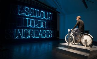 Pic 2 of 3 - Man on a bicycle in front of a neon sign reading "I SET OUT TO DO INCREASES"