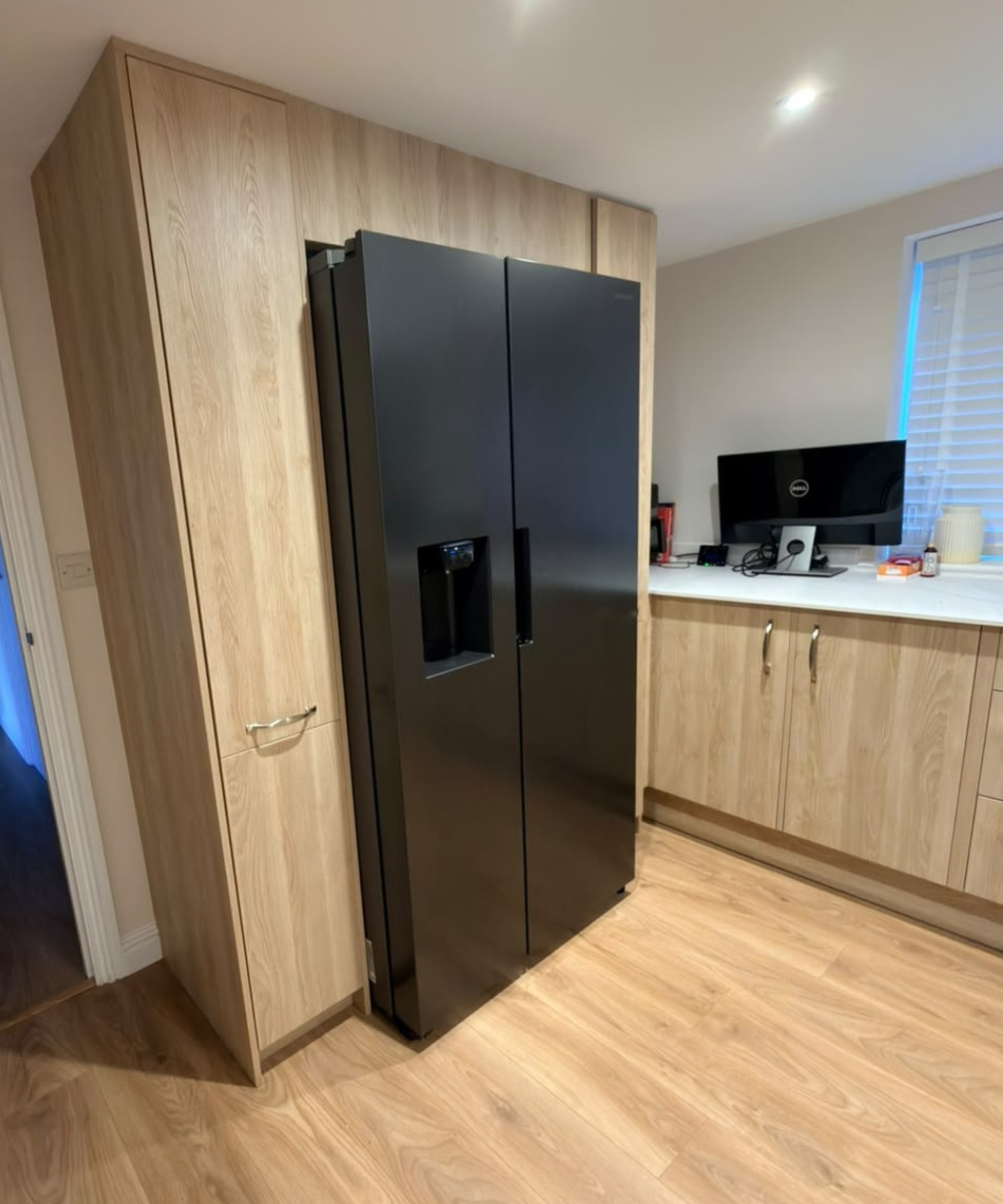 Black American-style fridge freezer in light oak tall units, next to a white worktop with a Dell monitor