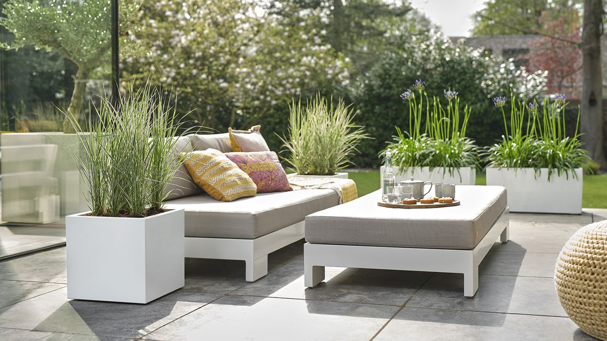 A contemporary patio with white geometric cpontainers with grasses and agapanthus next to a pale grey oudoor sofa and cofee table