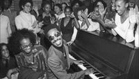 A black and white photo of Stevie Wonder at the piano, entertaining a dance theatre of Harlem students during a tour in New York, on 18th December 1976.