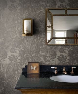 A bathroom with dark gray leafy wallpaper, a gold and marble wall sconce, a rectangular gold mirror, a black sink with a butterfly frame and brushed nickel faucets above the white sink