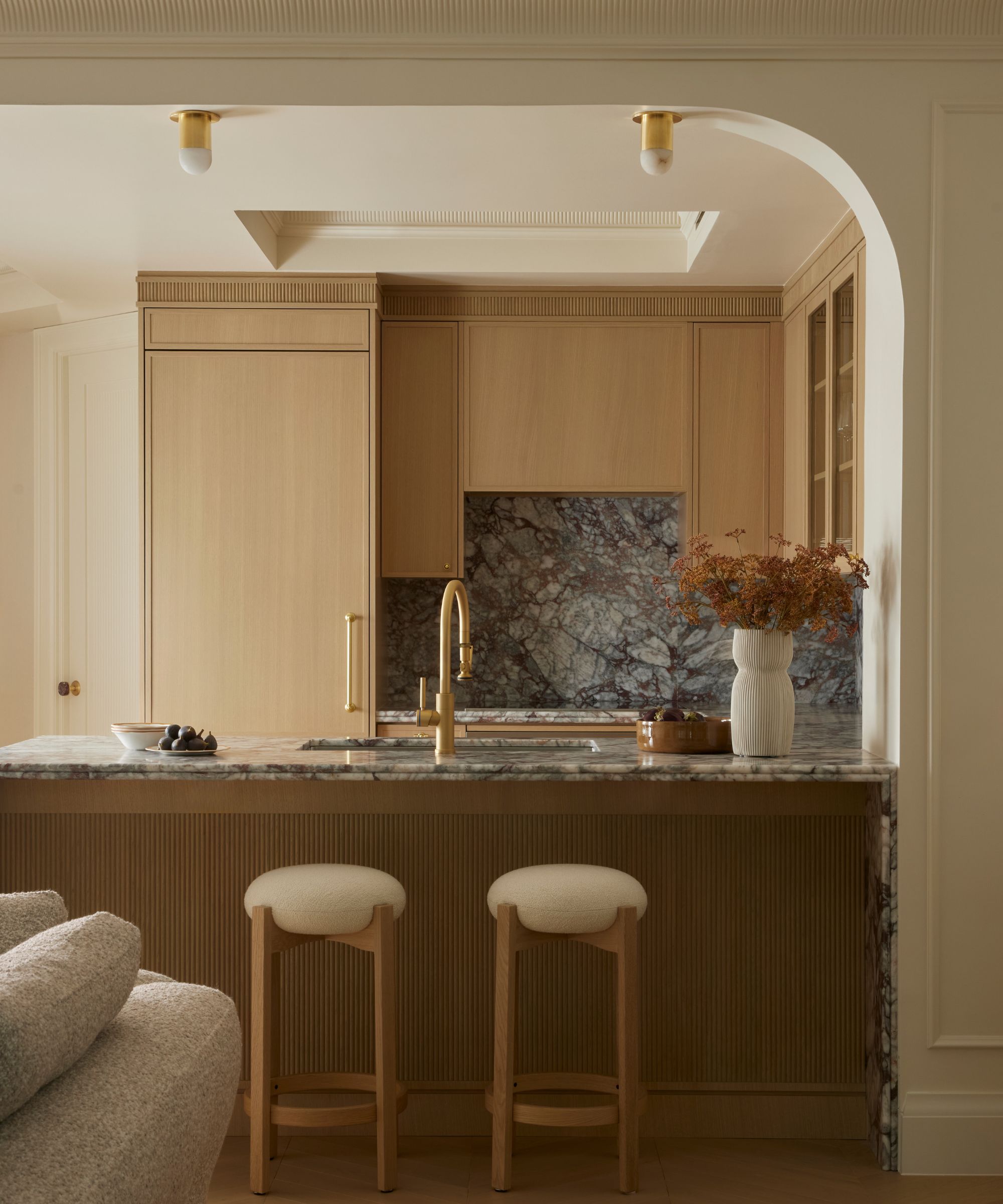 Modern kitchen bar with a dramatic purple-veined marble countertop and backsplash, golden faucet, light wood cabinets, and two cream-cushioned barstools.