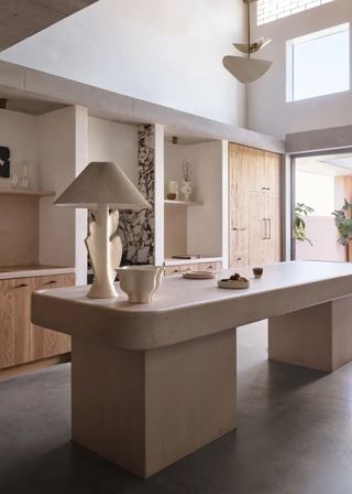 Kitchen cabinetry with a concrete island opposite it and wood cabinets behind. A lamp sits atop the island with a large bowl and plate.