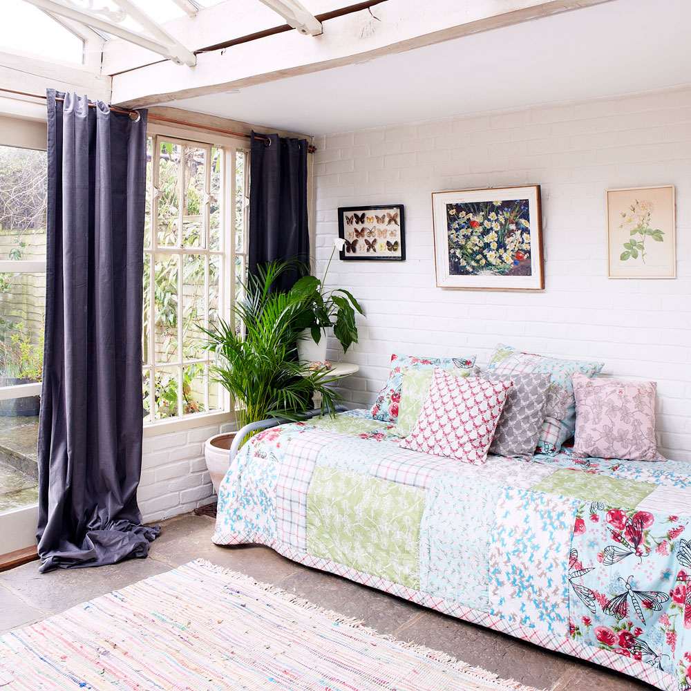 a day bed with a flower pattern duvet cover, in a conservatory with white brick walls, with long blue curtains in front of the window overlooking the outdoors patio