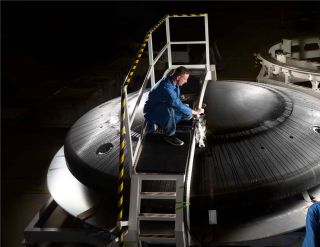 a man in blue jumpsuit facing right, rests his right hand on a low rail, while sitting on a over scaffolding arching over a black dome of a heat shield.