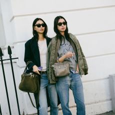 Models pose together for camera. One is wearing a black blazer, tee shirt, and jeans while carrying large handbag.The other friend is wearing a layered gray sweatshirt over jeans with a olive jacket on her shoulders with a asmall handbag. 