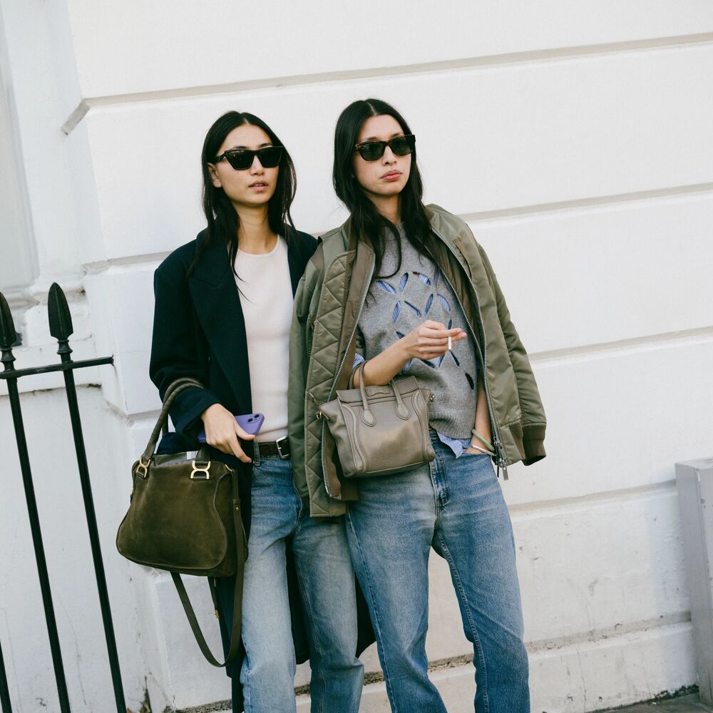 Models pose together for camera. One is wearing a black blazer, tee shirt, and jeans while carrying large handbag.The other friend is wearing a layered gray sweatshirt over jeans with a olive jacket on her shoulders with a asmall handbag. 
