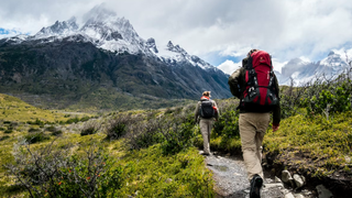 A pair of hikers are moving towards the mountains, they carry packs and are dressed for cooler weather in pants and jackets. 