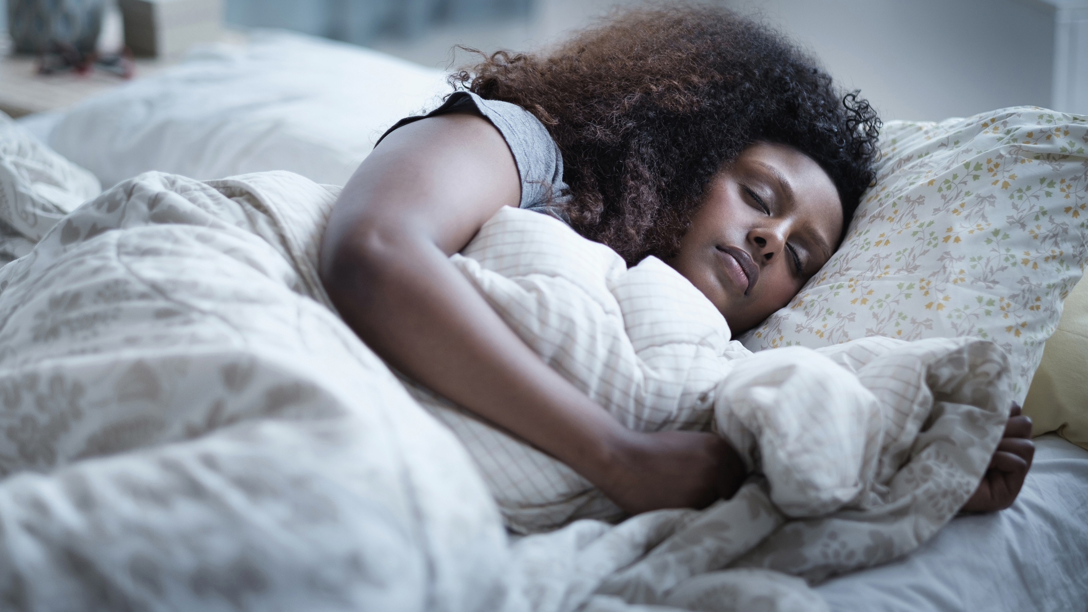 A woman with dark curly hair sleeps on a plush mattress and pillows