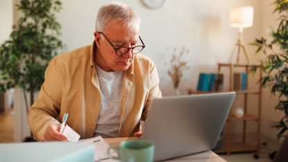 An older man concentrates while looking at his laptop screen and holding paperwork.