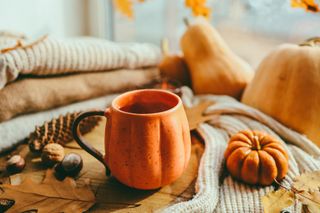 A cup of tea in a pumpkin shaped mug with a pumpkin on a wooden side table.