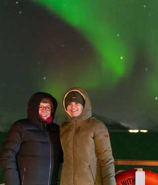 Two women wearing long coats and smiling with the northern lights behind.