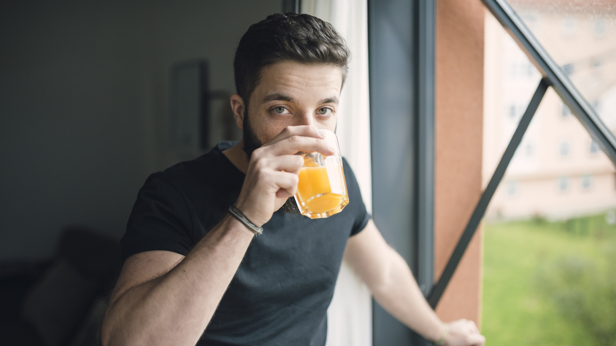 Man stands next to window drinking from glass and looks at the viewer