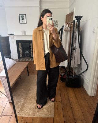 British fashion influencer Anna of The Anna Edit poses for mirror selfie wearing a camel jacket, beige crewneck sweater, brown leather snakeskin tote bag, black linen pants, and strappy black flat sandals