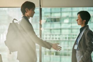 Shinichi Izumi (Masaki Suda) offers a hand to Jun-kyung (Lee Jung-hyun), while standing in a sunny glass-walled office, in the Parasyte: The Grey season 1 finale.
