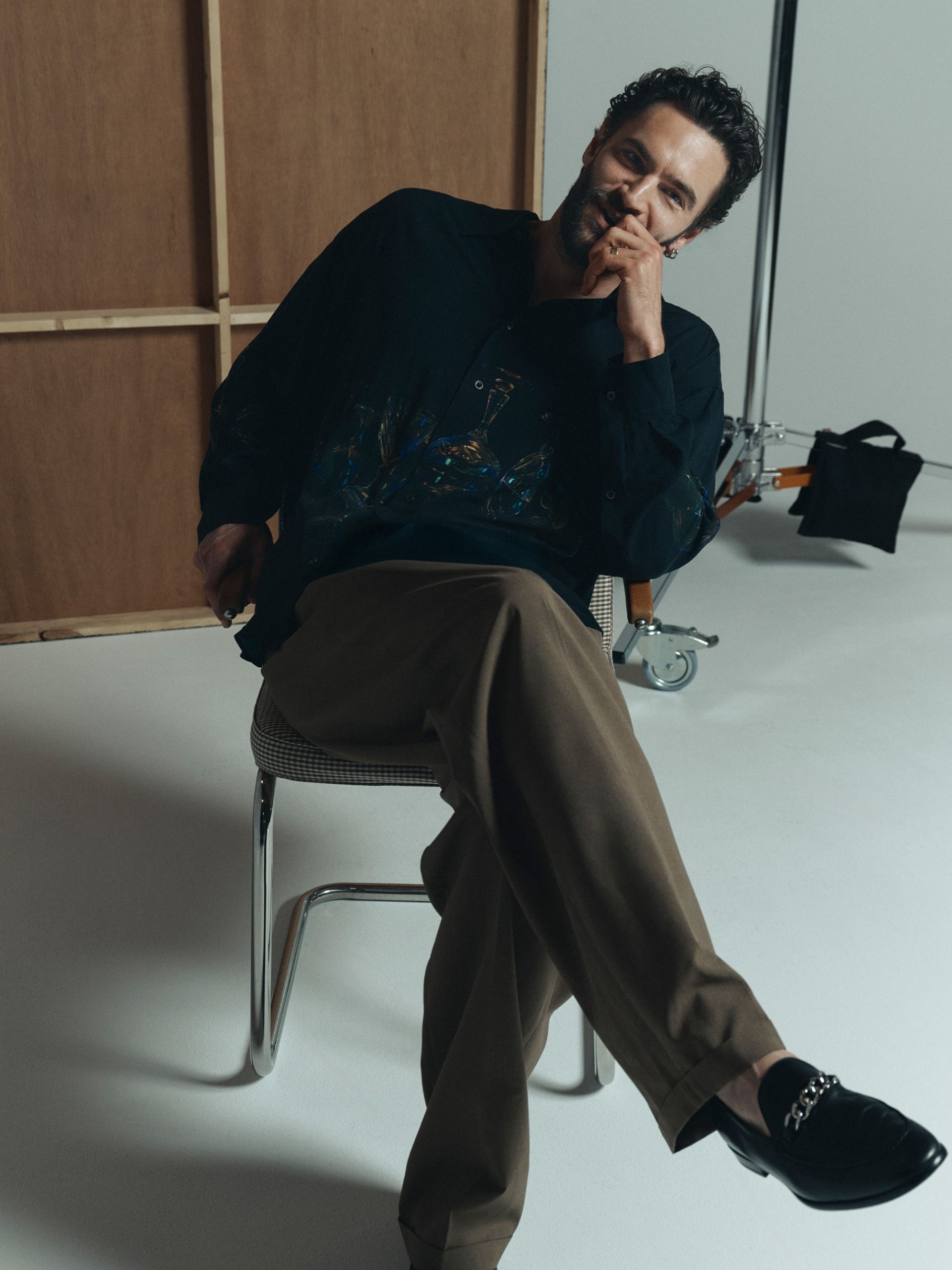 Actor Tom Bateman photographed by Graham Dunn. Shot in a studio, Bateman sits on a metal-frame chair wearing a dark blue button-down shirt with khaki trousers and loafers.