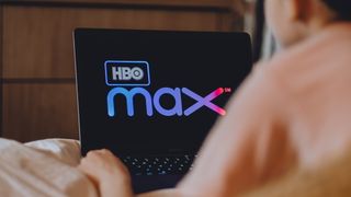 En person liggende i en seng med en bærbar PC med HBO Max-logoen synlig på skjermen