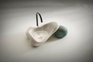 View from above of Daniel Arsham Kohler sink made of 3D printed white ceramic resting on an oxidised brass stone-shaped object, with water coming out of a minimalist black tap