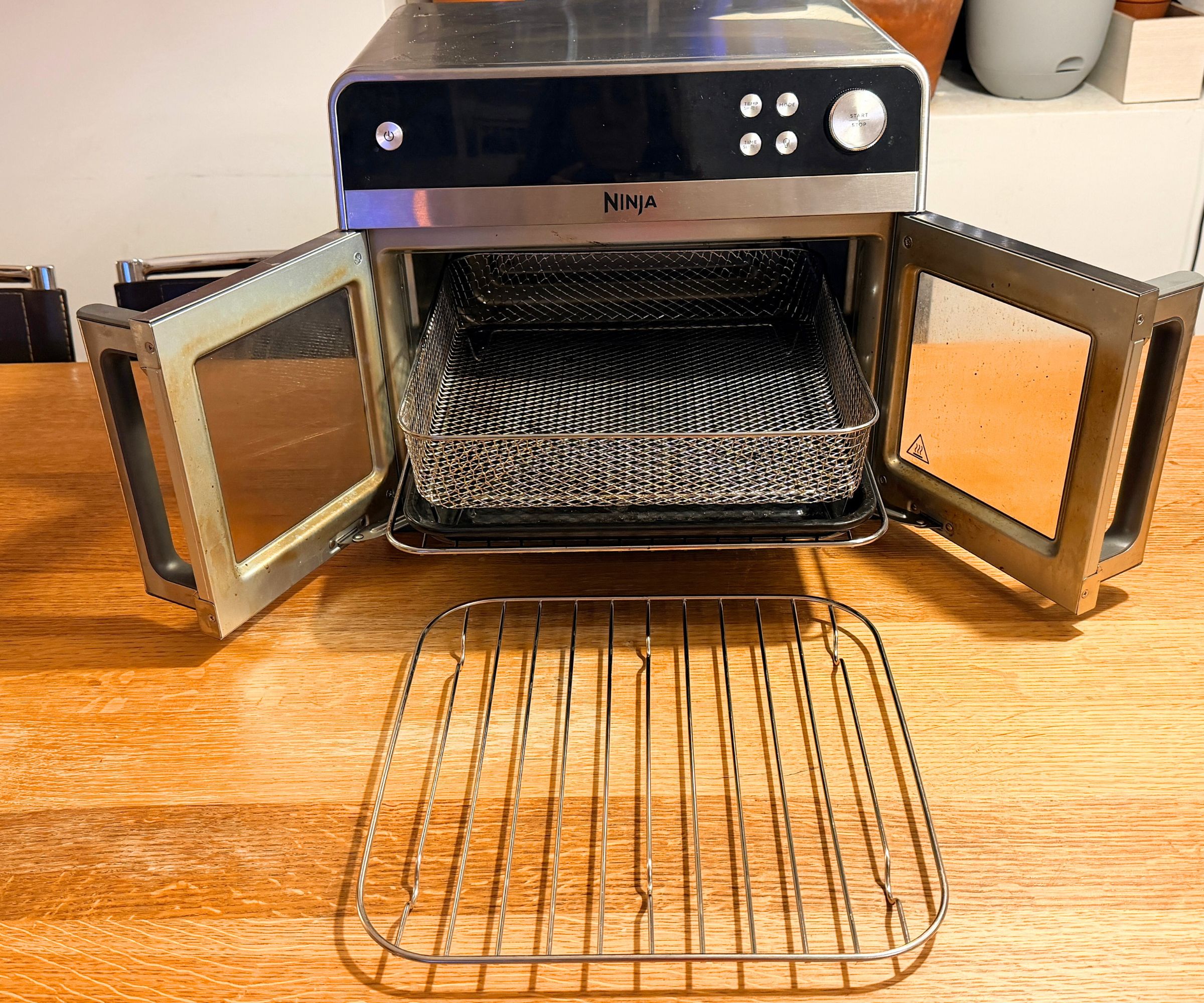 Ninja French Door Premier air fryer, open doors showing the metal tray and mesh basket inside, and a wire tray on the counter in front of it
