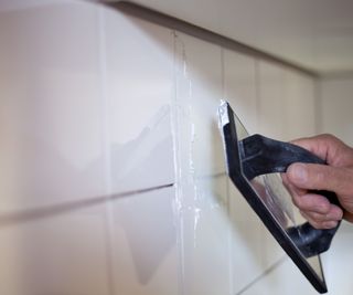 Someone using a black paddle board tool to apply grout to white tiles on a wall