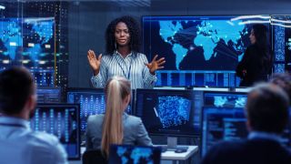 A woman giving a talk in front of colleagues inside a room filled with computer screens showing a map of the world