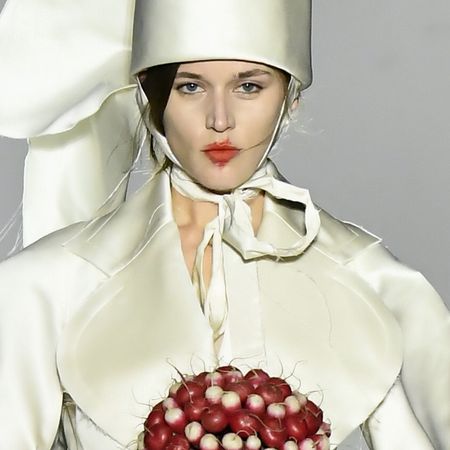 a model wears a tall ivory hat, an ivory blazer, and carries a radish bouquet.