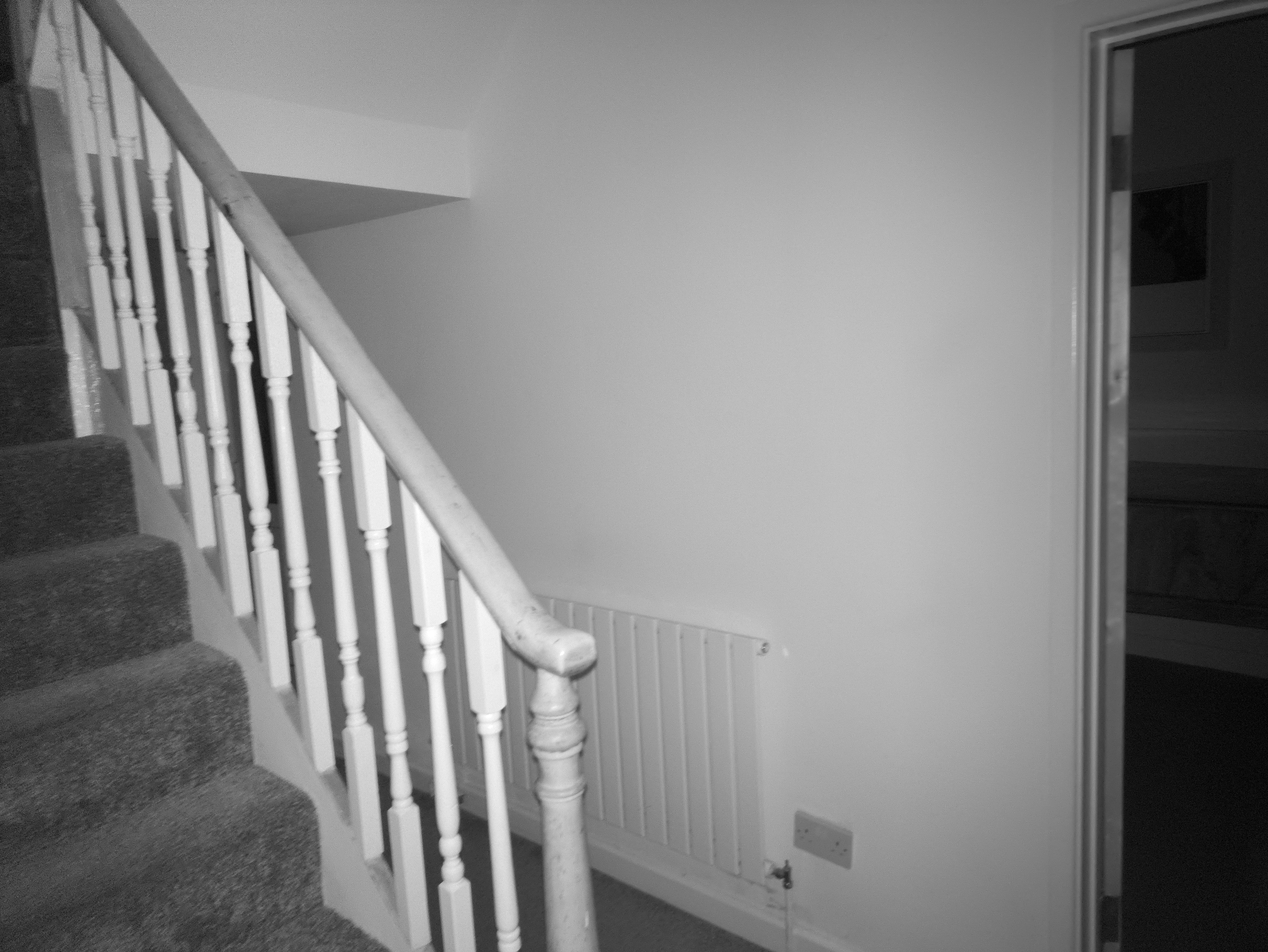 A view of an interior staircase with carpeted steps and a white painted wooden railing, alongside a wall-mounted radiator and a doorway into another room.