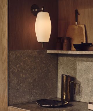 A moody kitchen nook with a frosted glass wall sconce, textured stone backsplash, warm wood cabinetry, and rustic decor, including wooden boards and a brass jug.