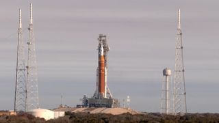 An orange rocket stands on a grey metal platform on a hill against a grey sky.