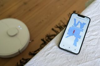 En mobil ligger på en möbelkant med den tillhörande appen för Dreame D10 Plus uppe på skärmen, med robotdammsugaren på golvet i bakgrunden.