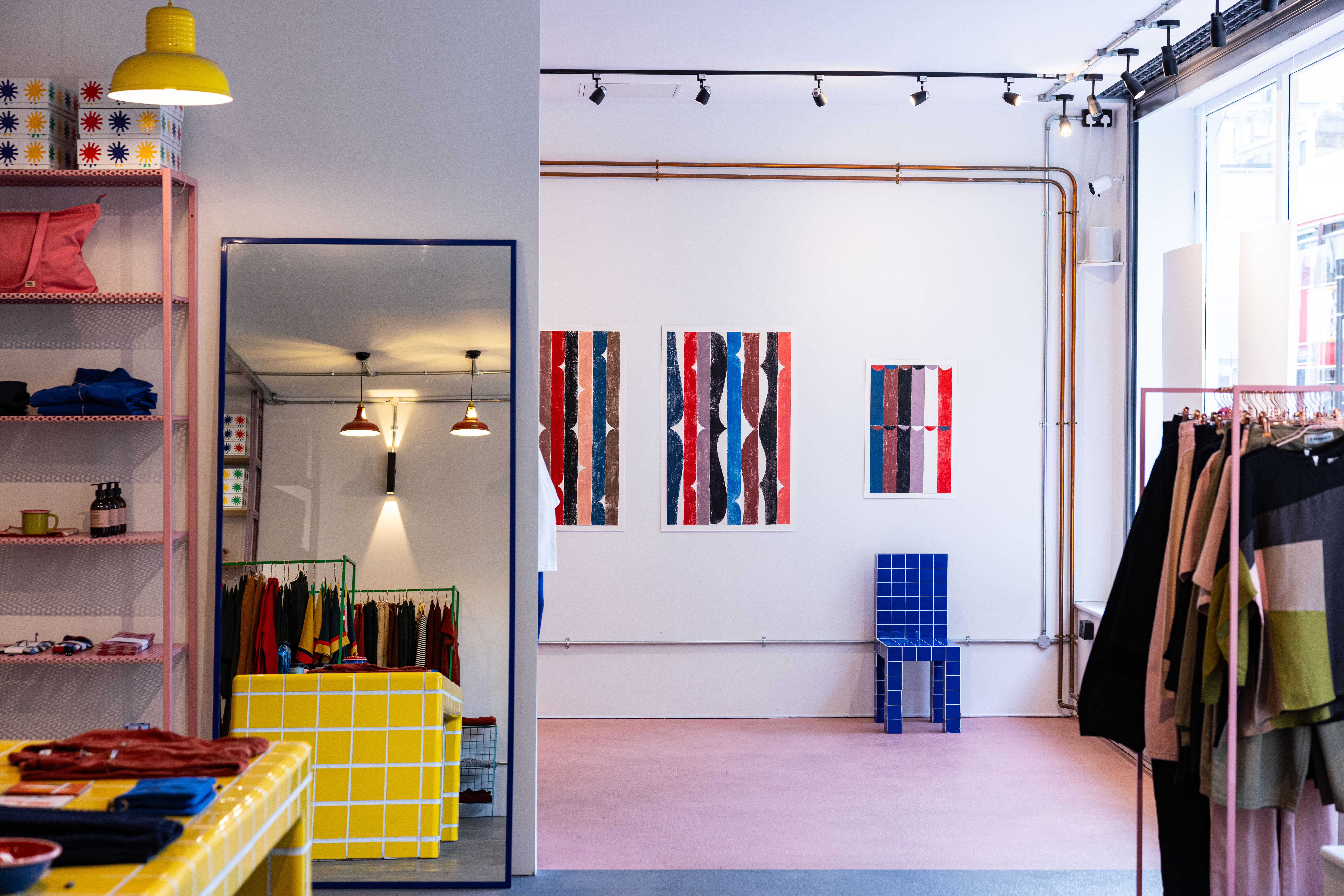 A clothing and design showroom hosts an exhibition of colorful, abstract prints now adorning its plain walls, immersed in colorful decor, and hosts the woman artist who designed them dressed in a double denim shirt and trousers outfit.
