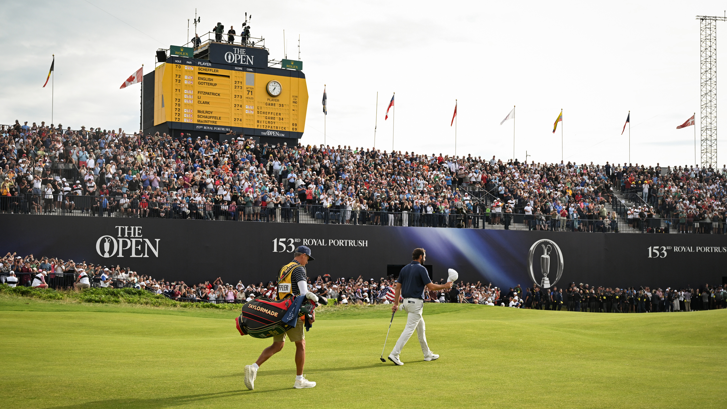 Scottie Scheffler waves his cap to the crowd as he approaches the 18th green at Royal Portrush. The iconic yellow scoreboard is in the background behind large galleries