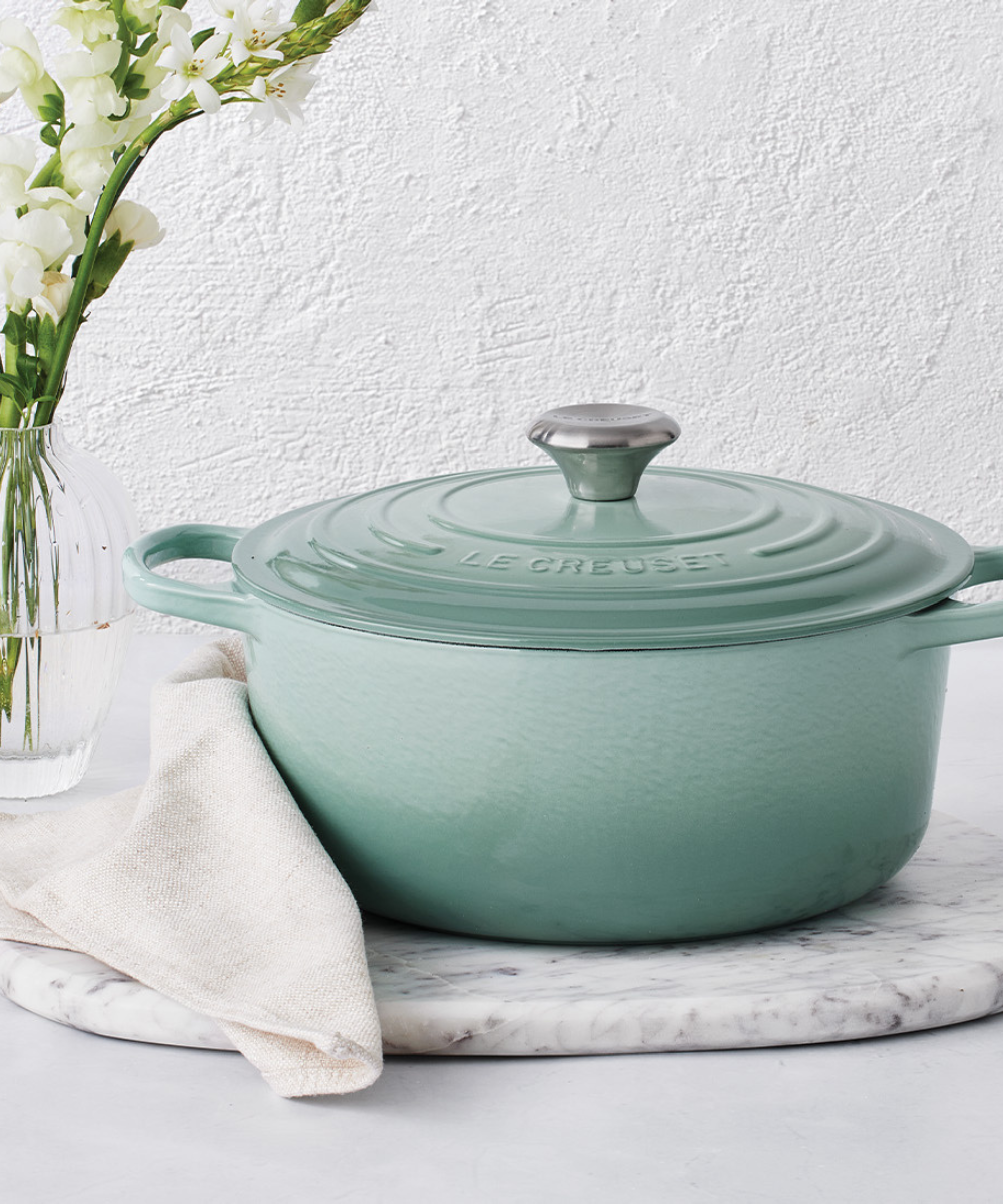 Le Creuset Signature Dutch oven in sage, against a white background, next to a linen napkin and white flowers in a glass vase