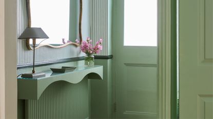 An entryway with wooden flooring, a light green arched front door, a green shelf above a green radiator with a mirror, and striped light green and white wallpaper on the upper walls.