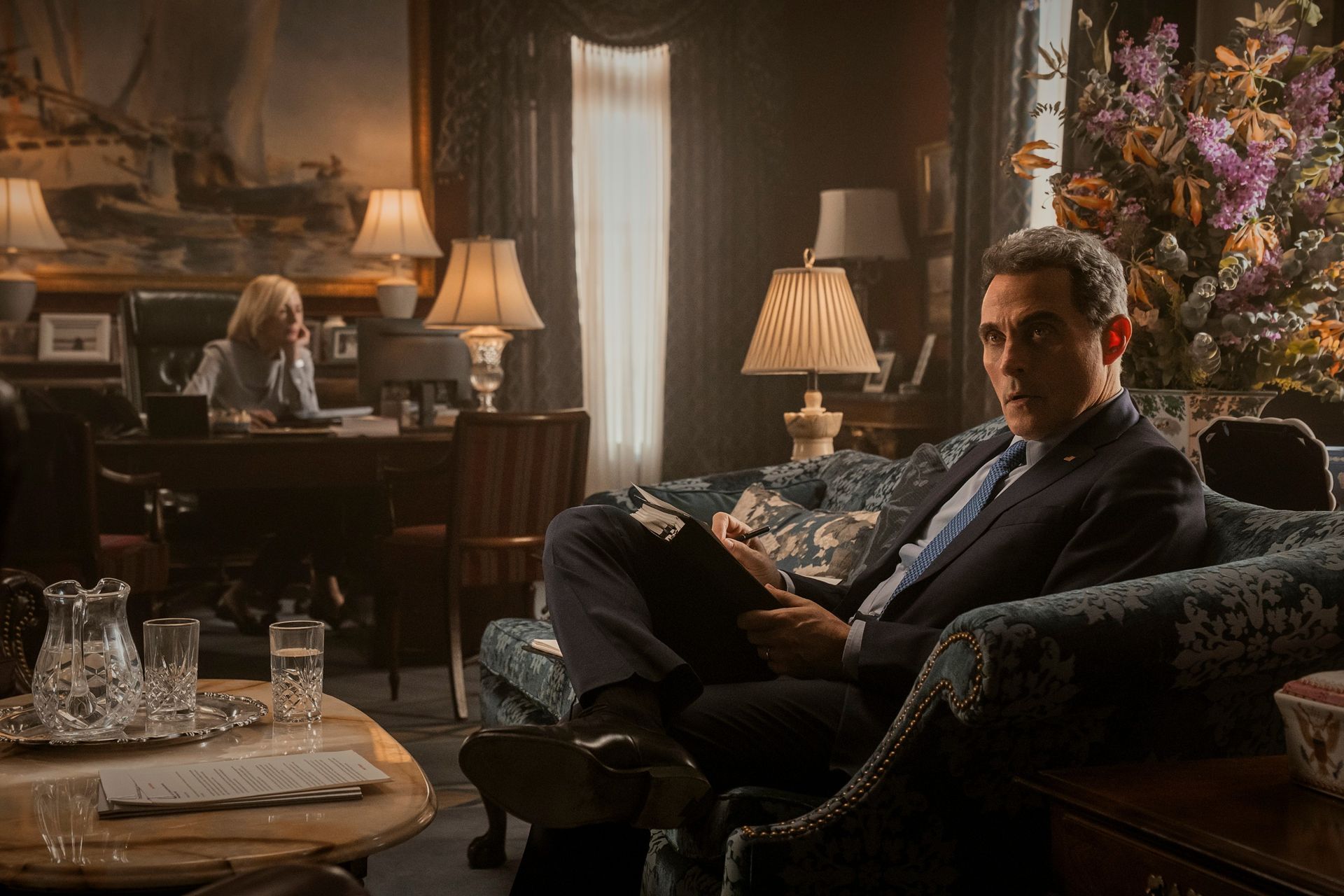 A woman (Allison Janney as Grace Penn) sits behind a desk in the background as a man (Rufus Sewell as Hal Wyler) sits on a loveseat in a grand home office, in 'The Diplomat' season 4.