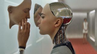 Still from the movie "Ex Machina." Close up of an android with a human face is touching a human face hanging on a wall.