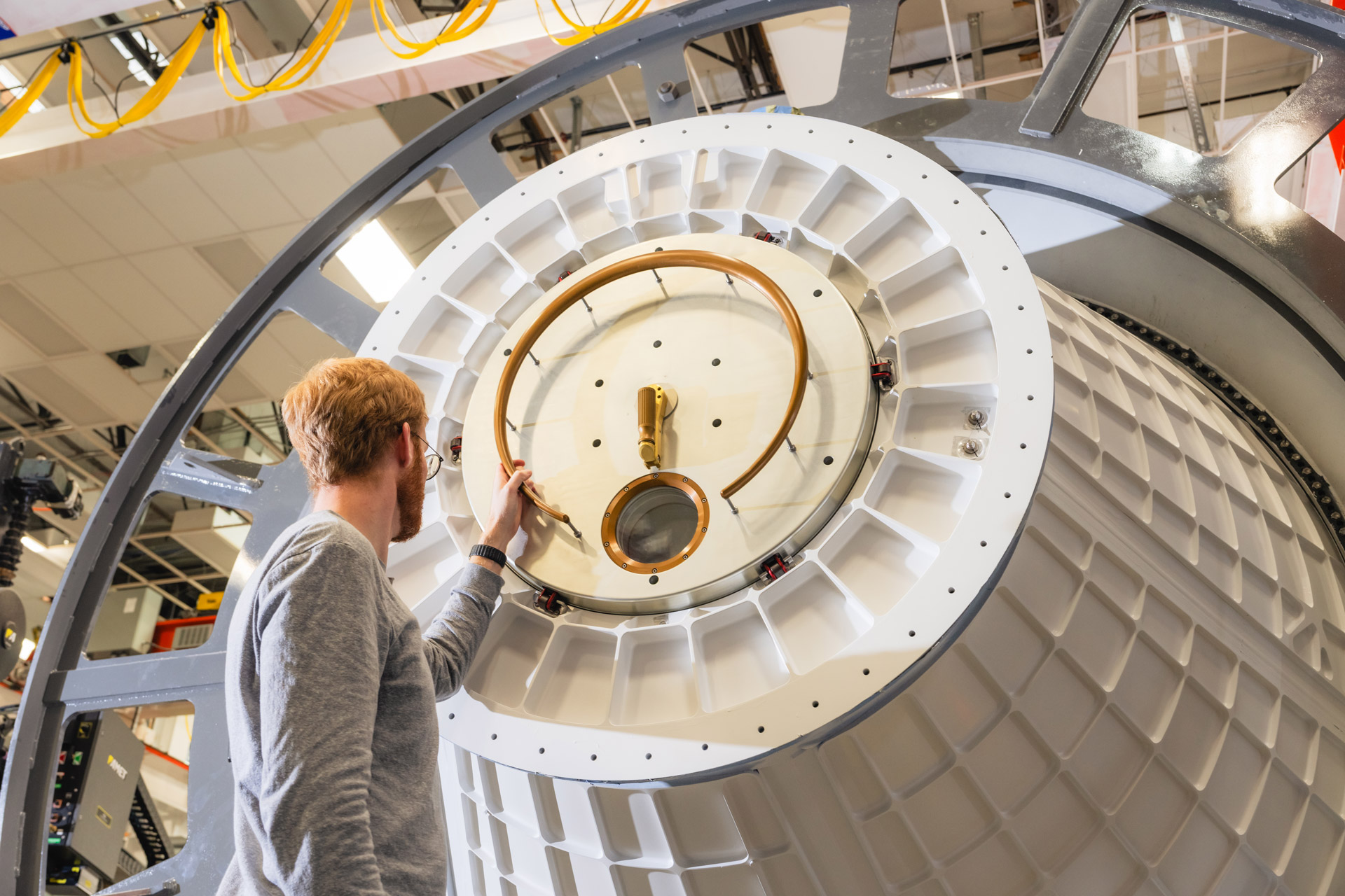 A man in a grey long-sleeve shirt and ginger hair faces away, looking up with his left hand holding the hatch at the end of a large white conical vessel with a gridded latus around its exterior.