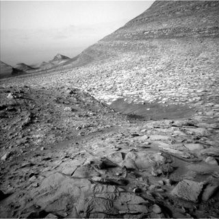 black and white view of the martian surface. cracked, rocky ground stretches ahead to peaking hills on the left horizon, the sky blocked on the right by a close, steep incline.