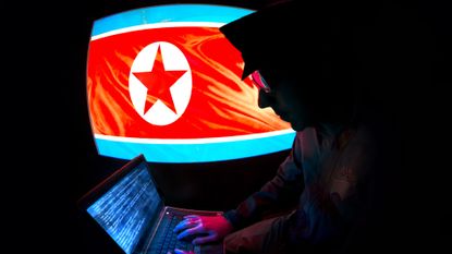 A North Korean IT worker photographed in side profile, silhouetted in the dark against a large monitor displaying the North Korean flag and looking at a laptop in their lap, on which code can be seen.