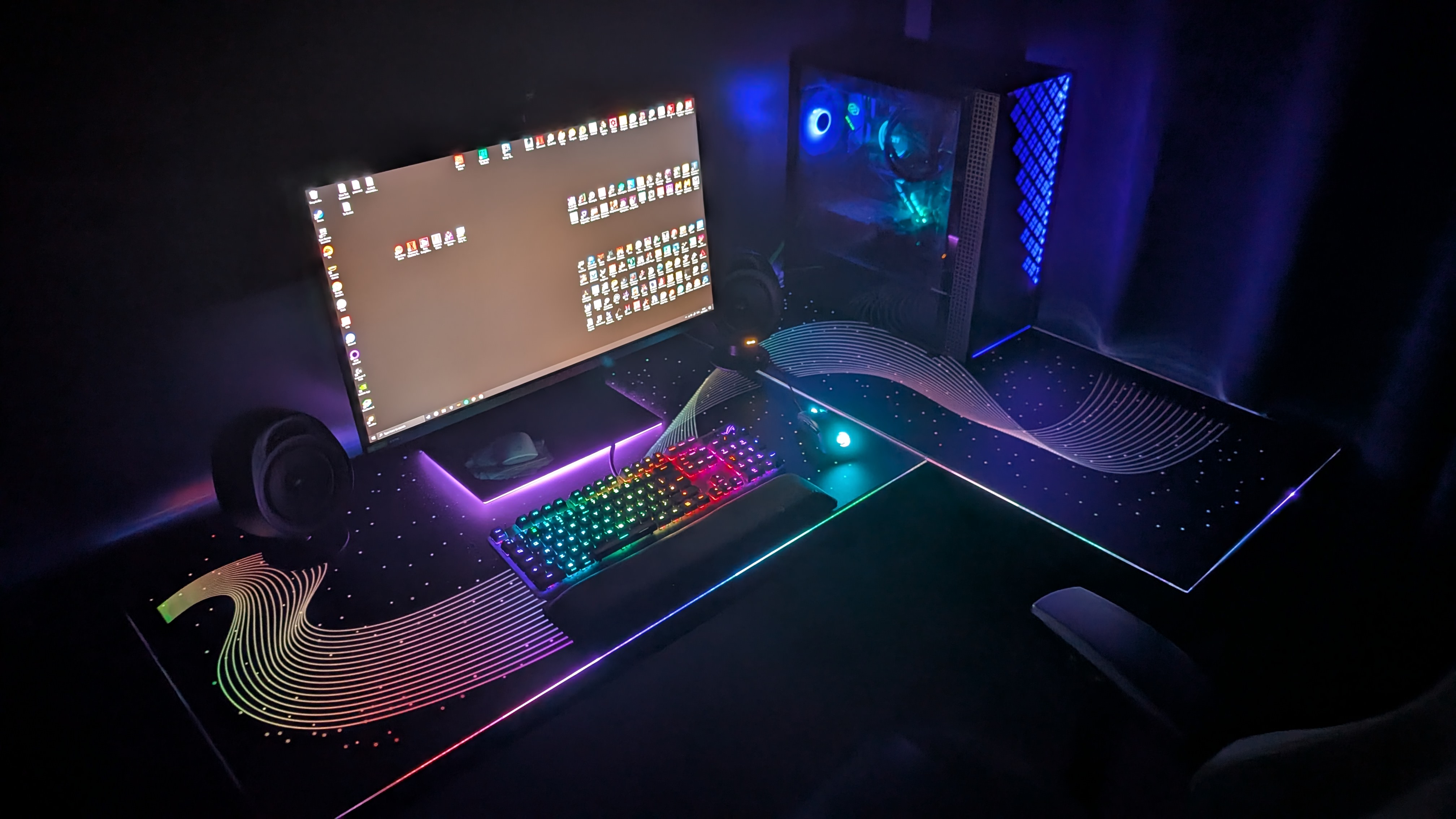 The Eureka Ergonomic GTG Wave gaming desk in a dark room with a PC setup on it, with all the RGB lights turned on to produce a rainbow wave pattern across the desktop.