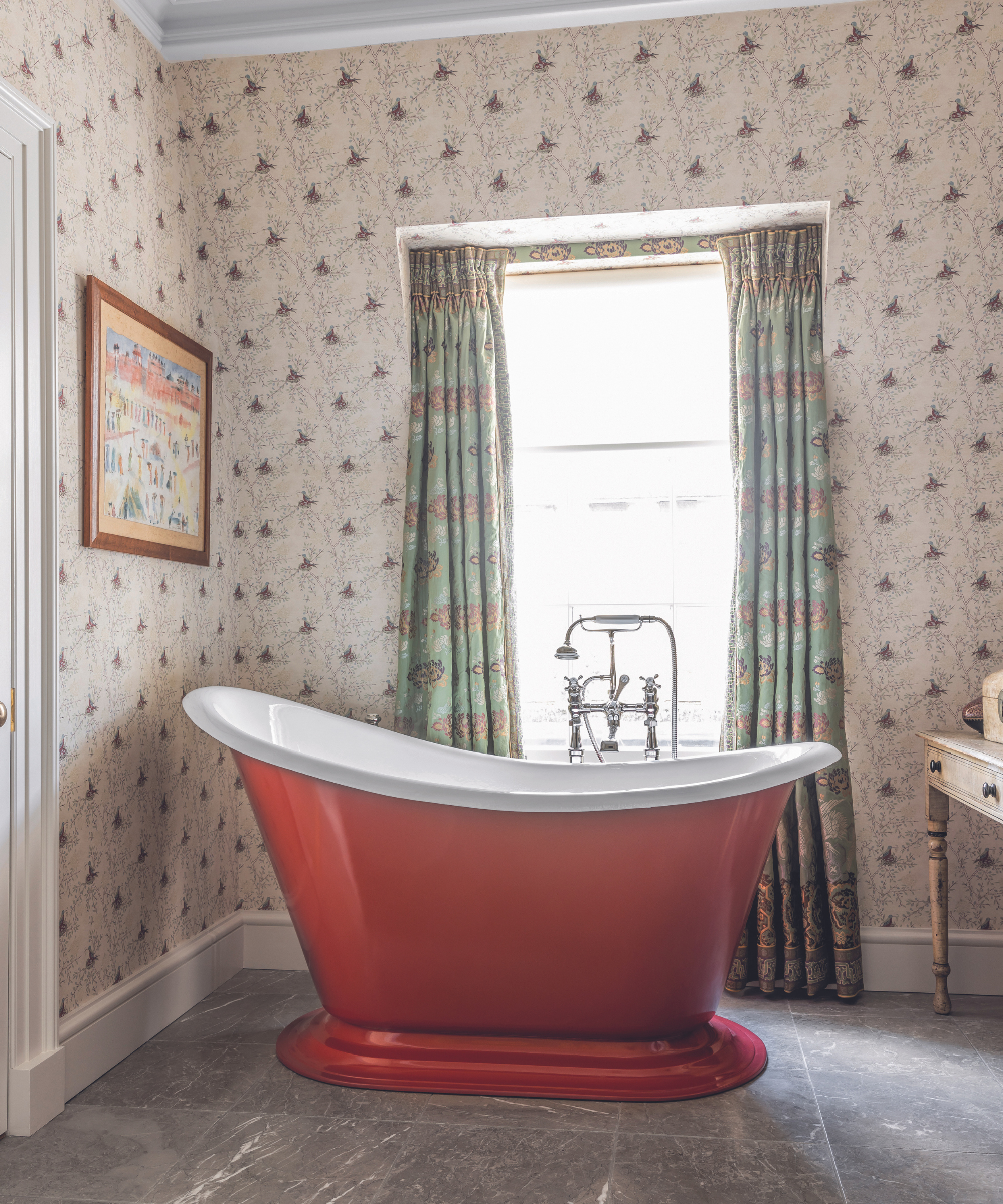 red freestanding bath in bathroom with grey stone floor and patterned wallpaper and curtains