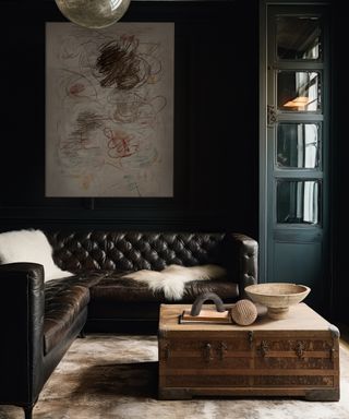 Living room with dark brown distressed leather sectional, trunk coffee table with vases, dark walls and large abstract artwork