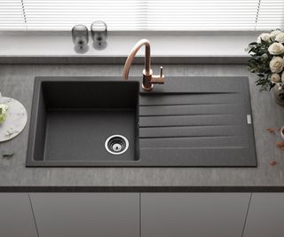 A modern kitchen sink setup featuring a matte black composite sink with an integrated draining board. The sink is paired with a rose gold single-lever tap, adding a warm metallic accent.