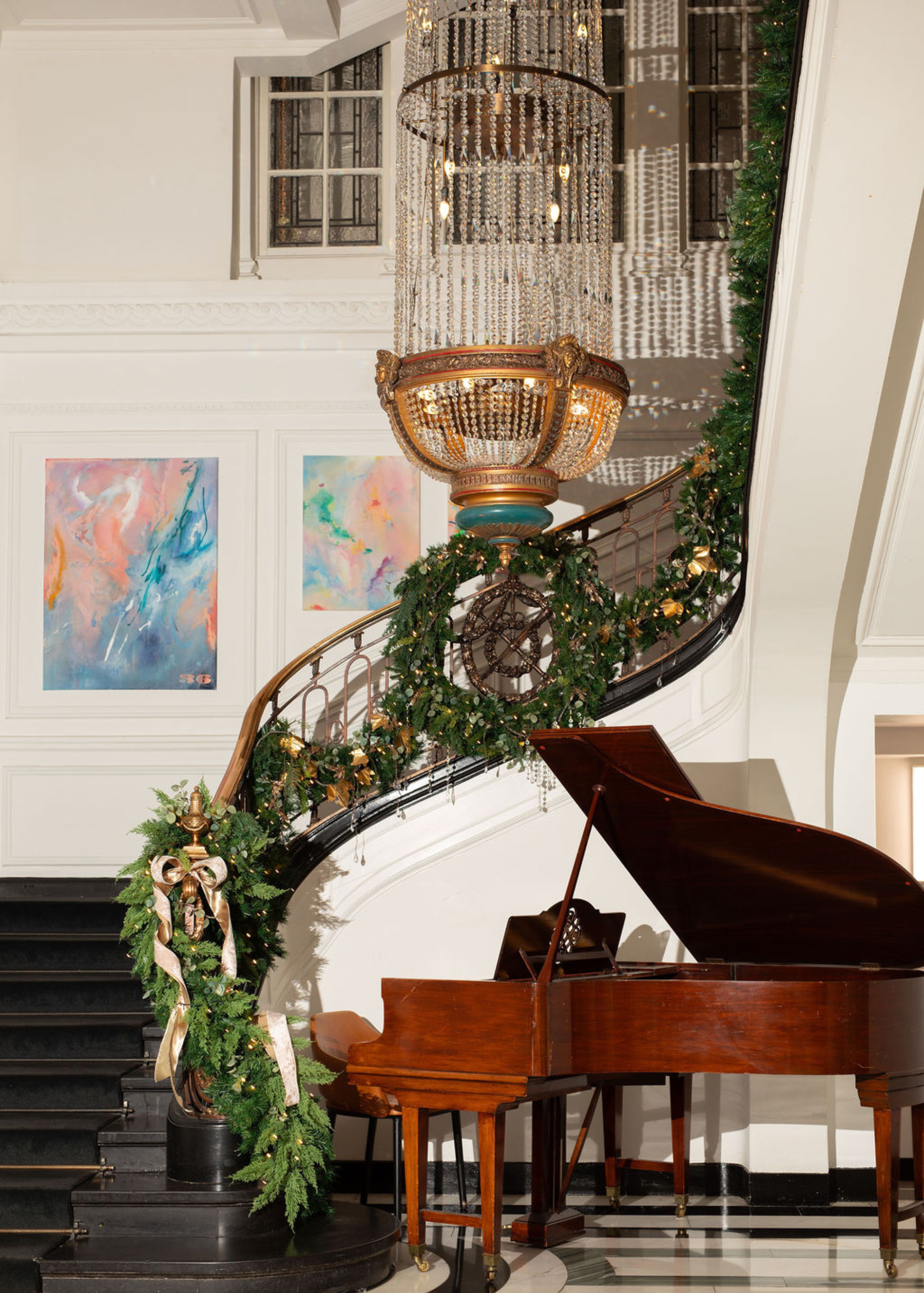 A large spiral staircase with a wreath and Christmas foliage wrapped around it. There is also a large chandelier hanging above a dark wood piano in a hotel lobby.
