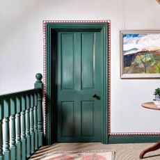 hallway with white walls and green doors and banister with red and white geometric border around the door and skirting border with natural carpet and artwork on the wall