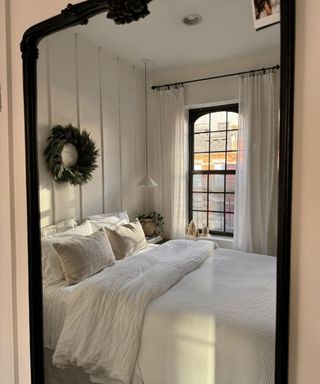 A dark ornate mirror reflecting a white bedroom with a white bed, window, wreath on panelled white wall behind bed